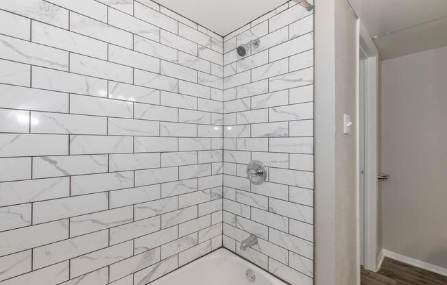 A clean bathroom featuring a shower area with white and gray marble-patterned tiles, a silver showerhead, and a bathtub. The walls are tiled in a grid pattern, and there is a closed door visible in the background, suggesting a modern aesthetic.