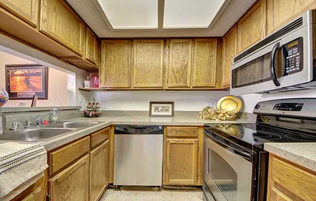 A cozy kitchen here at Summertree Place featuring warm wood cabinetry, durable countertops, a double-basin sink, built-in dishwasher, and a full appliance setup with generous cabinet storage and bright overhead lighting that creates a functional and welcoming cooking space.