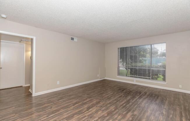 A spacious living room with light-colored walls and hardwood-style flooring. A large window allows natural light to fill the space, showcasing a view of greenery outside. An open doorway on the left leads to another room, creating an inviting and airy atmosphere.