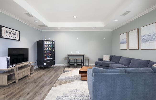 A cozy game room featuring a blue sectional couch, a wooden coffee table, and a small dining table with chairs. There's a flat-screen TV on a light wooden stand, and a black vending machine with snacks and drinks. The room has light gray walls and decorative artwork on the wall.