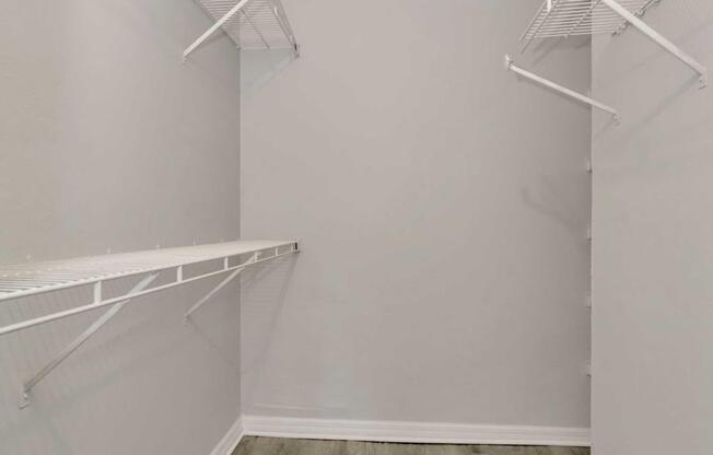 Empty closet with light gray walls and white wire shelving. Two shelves are mounted on opposite walls, and the floor is a light-colored wood or laminate. The space is well-lit and devoid of any items, providing a clean and minimalist appearance.
