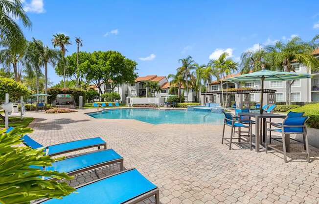 A pool surrounded by palm trees and a patio area with chairs and a table.