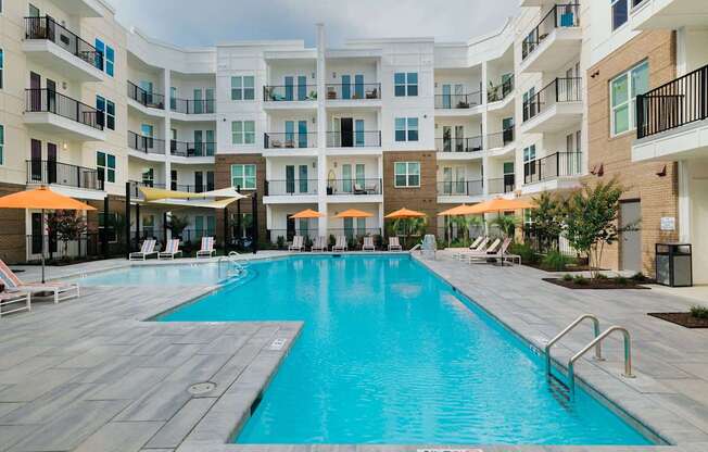 A large swimming pool in a residential building surrounded by lounge chairs and umbrellas.
