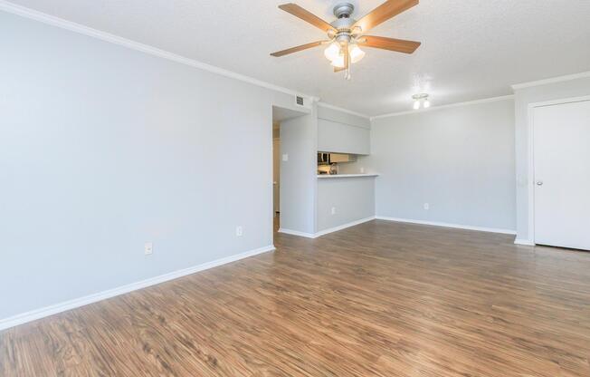 Spacious, empty room with light-colored walls and laminate flooring. A ceiling fan is installed in the center, and a doorway leads to a kitchen area with partial visibility. Natural light brightens the space, creating a calm and inviting atmosphere, ideal for interior design and decoration.