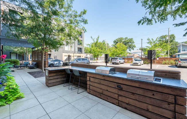 A wooden bar with a counter and chairs is in the foreground of a sunny outdoor patio.