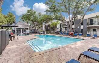 A pool surrounded by a brick patio and lounge chairs.