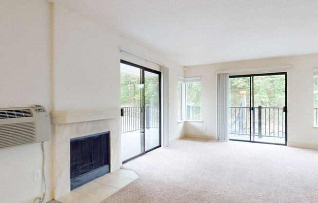 A spacious unfurnished living room here at Heritage Hills featuring soft neutral carpeting, a built-in fireplace with a tiled surround, and multiple sliding glass doors opening to private balconies. Large windows and glass doors bring in natural light and showcase wooded views, creating an open and airy space ideal for flexible furniture layouts.