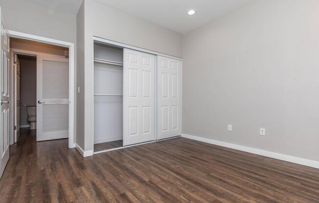 A spacious, empty room with light gray walls and hardwood flooring. It features a sliding closet with white doors, a doorway leading to another room, and a ceiling light. There are no furnishings or decorations, creating a clean and modern look.