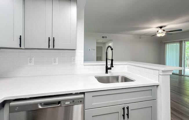Modern kitchen featuring gray cabinets, a white countertop, a stainless steel dishwasher, and a sleek black faucet over a stainless steel sink. The background shows a neutral-colored living area with a ceiling fan and sliding glass doors, providing a bright, airy atmosphere.