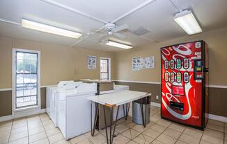 A bright and clean laundry room featuring two white washing machines, a folding table, and a red Coca-Cola vending machine. There are two windows allowing natural light in, and informational posters are displayed on the walls.
