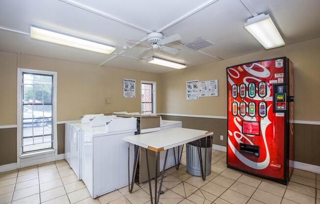 A bright and clean laundry room featuring two white washing machines, a folding table, and a red Coca-Cola vending machine. There are two windows allowing natural light in, and informational posters are displayed on the walls.