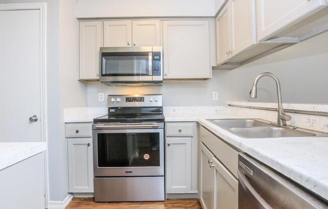 A modern kitchen featuring light gray cabinets, a stainless steel oven and microwave, and a double sink. The countertops are a speckled white with gray marbling. The kitchen layout is compact and includes a doorway leading to another room. The flooring is a warm wood laminate.