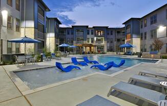 Modern apartment complex pool area at dusk, featuring a clear swimming pool surrounded by lounge chairs, blue umbrellas, and stylish architecture. The buildings have a contemporary design with large windows, and the scene is illuminated by soft lighting, creating a relaxing atmosphere.