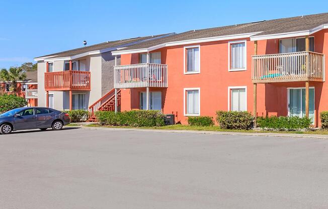 A photo of a two-story apartment building featuring orange and gray facades. Each unit has a balcony with railings, and some windows are open. In the foreground, there is a parking lot with a gray car parked, and well-maintained shrubs line the building, indicating a clear sunny day.