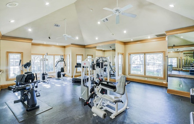 Fitness center at Saxony at Chase Oaks Apartments in Dallas, TX, featuring modern exercise equipment, a mirrored wall, vaulted ceilings with ceiling fans, and large windows letting in natural light.