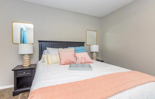A cozy bedroom featuring a dark wood bed with white and pastel-colored bedding. Two stylish bedside lamps flank the bed, and there are two mirrors on the walls. A gray tray with a glass is placed on the bed, alongside decorative pillows in soft hues. The walls are painted a neutral color, enhancing the inviting atmosphere.