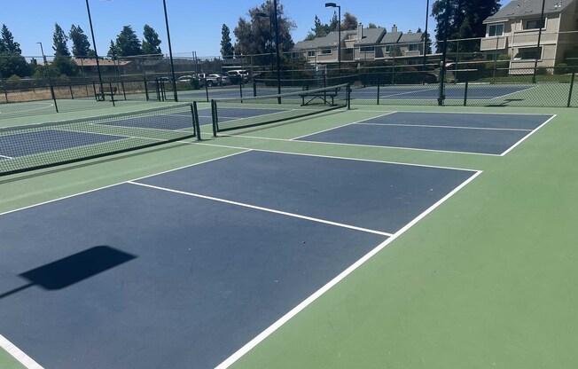 Empty outdoor tennis courts under a clear blue sky. Several courts are visible, with clear markings and a green surface. In the background, there are trees and residential buildings. Shadows from the net posts cast on the ground. The scene conveys a peaceful, sunny day, ideal for playing tennis.