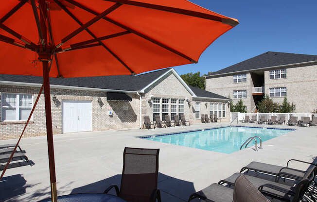 A large orange umbrella is shading a table and chairs by a pool.