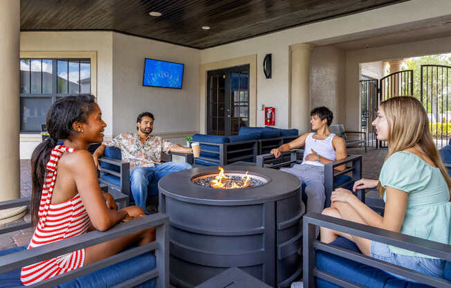 A group of people are sitting around a fire pit in a room with a TV screen on the wall.