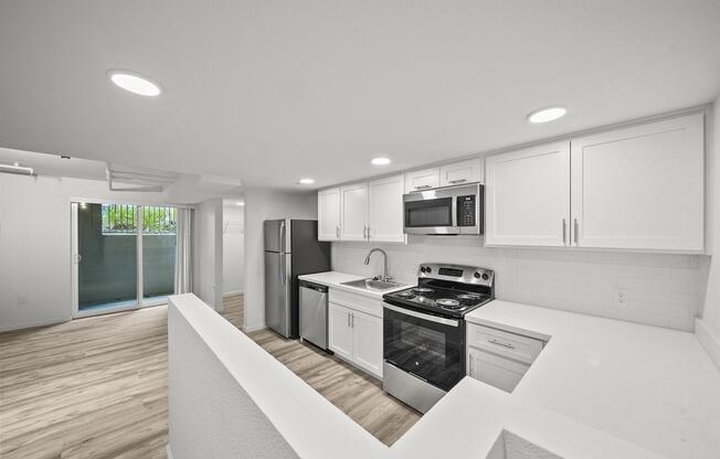 Modern kitchen featuring white cabinets, stainless steel appliances, a gas stove, and a large sink. The space includes a refrigerator and is open to a living area with light-colored wood flooring. Large windows in the background provide natural light, leading to a balcony.