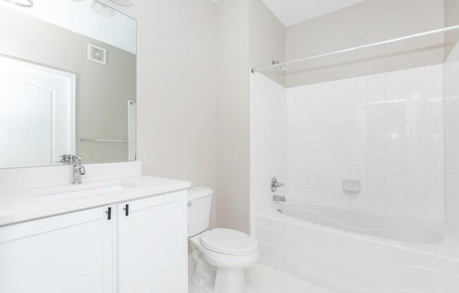 A clean, modern bathroom featuring a white bathtub and shower combo, a white toilet, and a vanity with a sink. The walls are a light beige color, and there is a large mirror above the sink. The overall atmosphere is bright and minimalist.