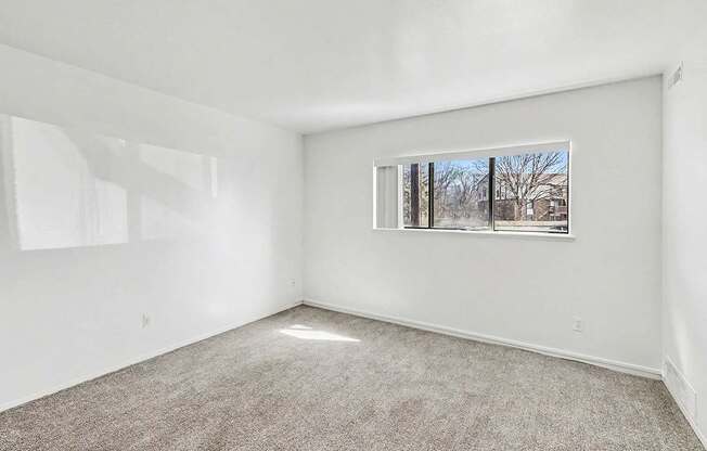 A garden style living room with a carpeted floor and a window showing a view of trees outside at Trappers Cove Apartments, Lansing