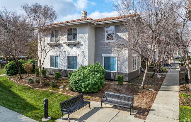 A house with a grey and white exterior is surrounded by a well-kept lawn and a sidewalk with two benches at Cornerstone at Gale Ranch Apartments, California, 94582