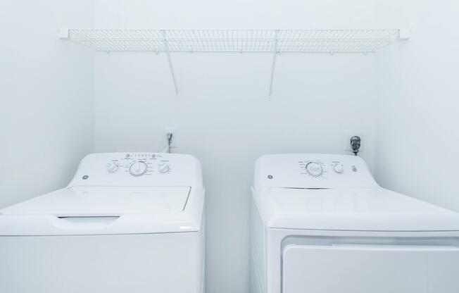 A clean and modern laundry room featuring a white washing machine and a white dryer side by side. Above them, a simple wire shelf is mounted on the wall for storage. The walls are painted white, creating a bright and spacious atmosphere.