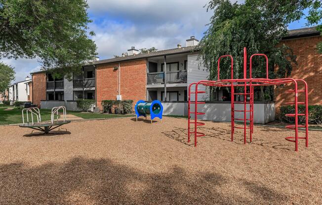 Playground area featuring a red jungle gym, blue tunnel slide, and a roundabout, surrounded by gravel and residential buildings with greenery.