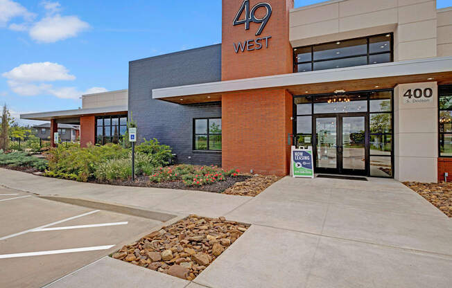 Modern building exterior with "49 West" sign, large glass entrance, and red brick facade. "Now Leasing" sign outside. Clear blue sky and greenery.
