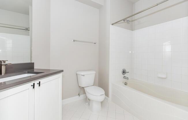 A clean and modern bathroom featuring a white bathtub, a shower curtain rod, a toilet, a sink with a dark countertop, and a large wall mirror. The walls are a light neutral color, and the flooring is tiled in a white pattern. The space is bright and uncluttered.
