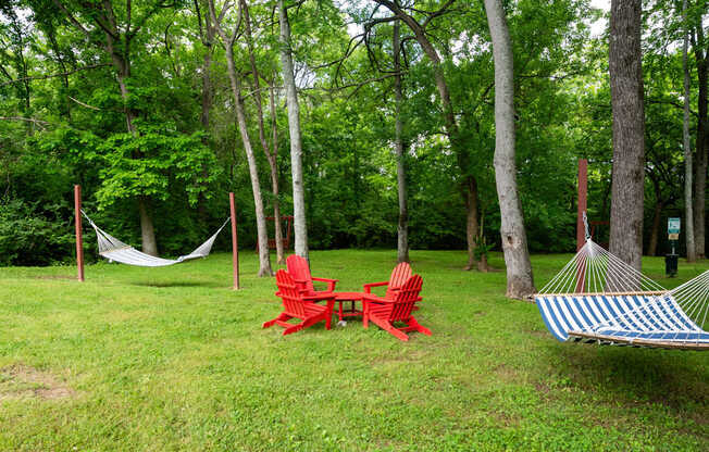 A red chair and a blue and white striped hammock are set up in a grassy area with trees in the background at the Retreat at Indian Lake Apartments.
