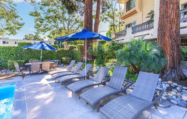 Lounge chairs with blue umbrellas near a swimming pool, surrounded by lush greenery and trees. A dining set is visible in the background, creating a relaxing outdoor atmosphere. The setting is well-lit, showcasing a bright and inviting environment.