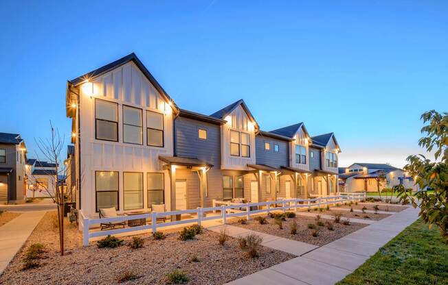 A row of houses with lit up windows and a white fence in front.
