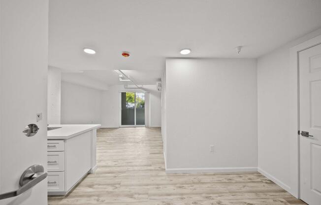 Bright, modern interior of an apartment featuring an open floor plan. The space includes a kitchen area with white cabinetry and a door leading to a patio or balcony. The walls are painted white, and flooring is light-colored wood. Ample natural light filters in through the sliding glass doors.