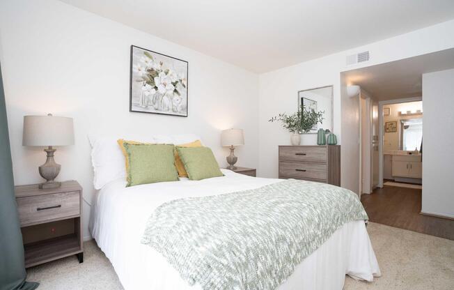 A cozy bedroom featuring a bed with green and white bedding, two bedside lamps, and a decorative painting on the wall. There is a dresser with plants and a mirror, and a bathroom is visible in the background. The room has light-colored walls and carpet, creating a warm and inviting atmosphere.