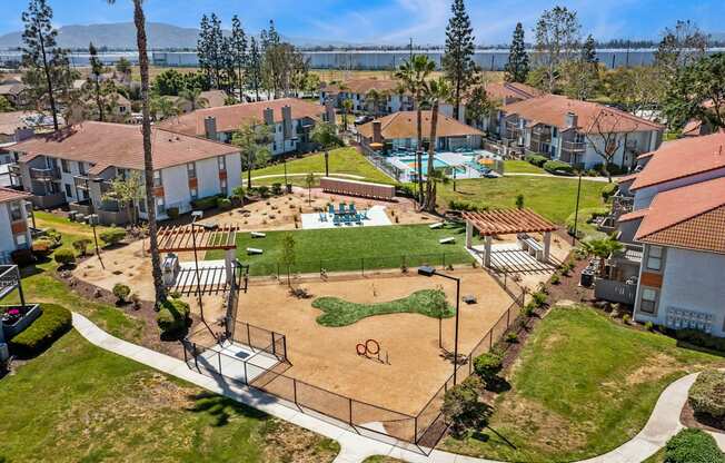 Dog-Friendly Apartments in Moreno Valley, CA - Ardella on Chagall - Aerial View of Property Grounds Showcasing Aparment Buildings, Fenced Dog Park, BBQ Area with Grilling Stations, Cornhole Playing Field, and Pool Area.