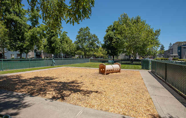 A dog park with a large brown barrel in the middle of a wood chip area at Autumn Oaks Apartments, Roseville, CA, 95678