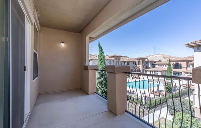 A spacious balcony with a view of a swimming pool and landscaped area. Tall green trees are visible, along with several buildings in the background. The sky is clear and blue, indicating a sunny day. The balcony features a railing and a light fixture on the wall.