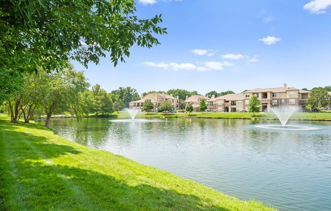A serene lake with a fountain in the middle surrounded by lush greenery and apartment buildings in the distance.