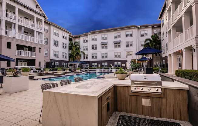 A poolside grill is in front of a pool and apartment buildings.