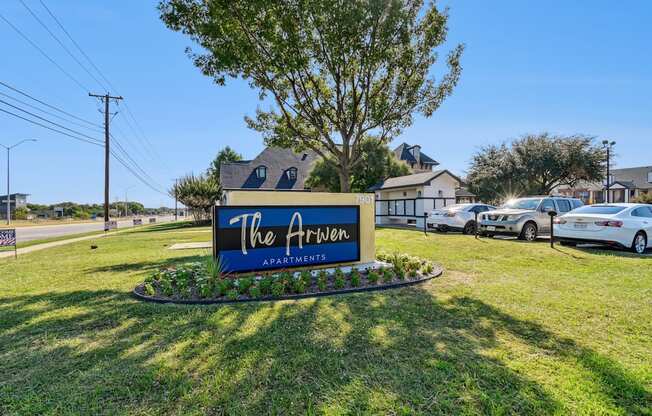 A sign for "The Arrow Apartments" is displayed in a grassy area with a tree and parked cars in the background.