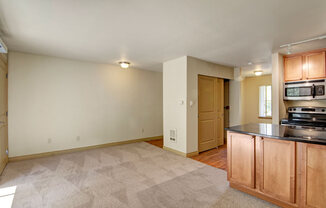 A spacious open living area here at Asbury Park featuring soft carpeted flooring, neutral walls, and a wide layout that flows into a functional kitchen with warm wood cabinetry, a dark countertop, and modern appliances, while ceiling lighting and a nearby window provide even illumination and a comfortable, practical atmosphere.