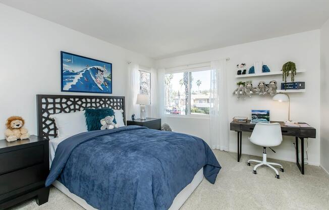 A cozy bedroom featuring a blue bedspread, a dark wood headboard, and light-colored walls. A window allows natural light, revealing palm trees outside. There's a small desk with a chair, shelves with decorative items, and a plush lion toy on the bedside table. The overall decor is simple and inviting.