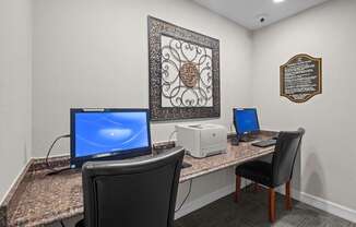 A room with a desk, two chairs, a monitor, a printer, and a framed artwork on the wall.