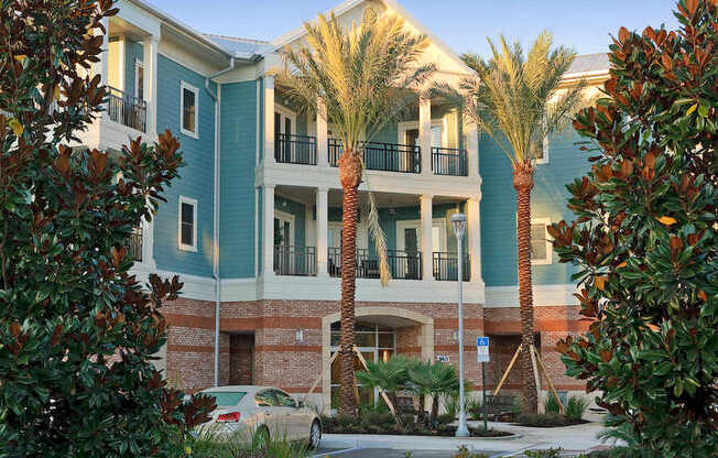 An exterior view of a 3-story apartment building at The Flats at Tioga Town Center with patios or balconies and well-maintained palm trees near resident parking.