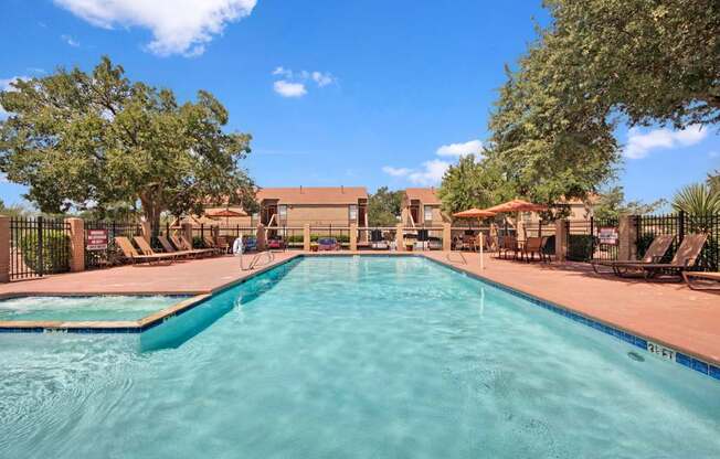 A spacious outdoor swimming pool here at Trinity Place with clear blue water and wide entry steps. Lounge chairs line the surrounding deck, with patio tables and umbrellas offering shaded seating. A fenced layout and mature trees create a private, relaxing atmosphere for residents to enjoy sunny days.