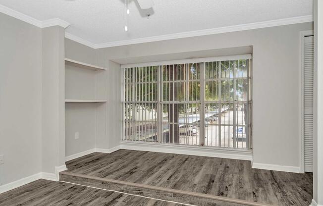 A well-lit room featuring large windows with blinds, allowing natural light to enter. The flooring is a modern wood style, and there is a small shelf along one wall. Outside the windows, a view of trees and a street is visible. The overall decor is neutral and contemporary.