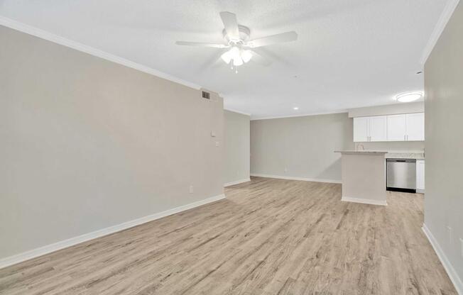A spacious, empty room featuring light-colored walls, a ceiling fan, and hardwood-style flooring. In the background, there is a kitchen area with white cabinets and stainless steel appliances. The open layout gives a modern feel with ample natural light.