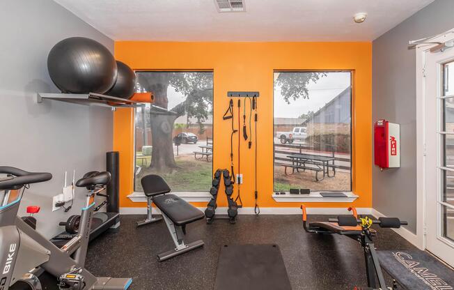 A bright and modern fitness room featuring various exercise equipment, including a stationary bike, weights, resistance bands, and a workout mat. Large windows provide natural light and a view outside, while an orange accent wall adds a pop of color. The space is neatly organized and conducive to workouts.